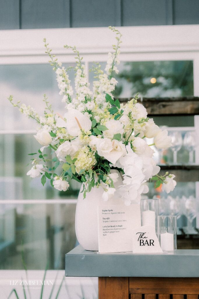 White floral arrangement in a vase on a table next to a sign labeled "The Bar" and a menu. Glasses are seen in the background.