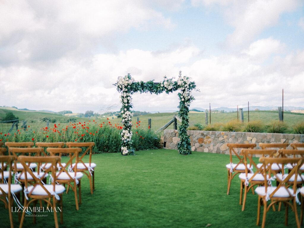 Outdoor wedding ceremony setup with wooden chairs facing a floral arch. Green grass and open sky in the background.