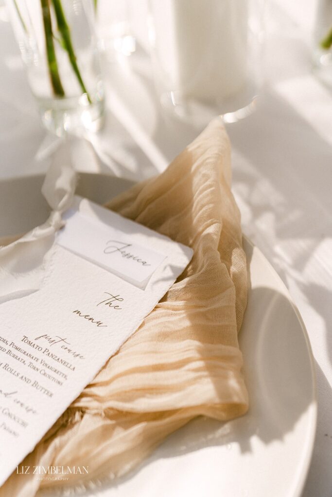 A beige napkin and menu set on a white plate with sunlight casting shadows.