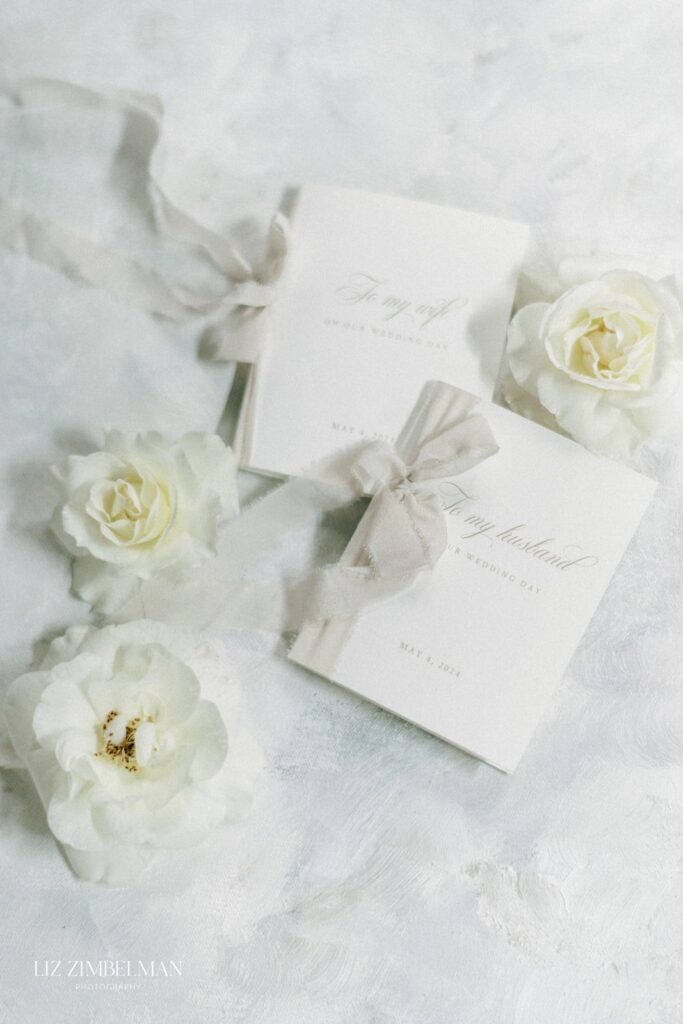 Two white wedding vow booklets with gray ribbons surrounded by white roses on a textured white surface.