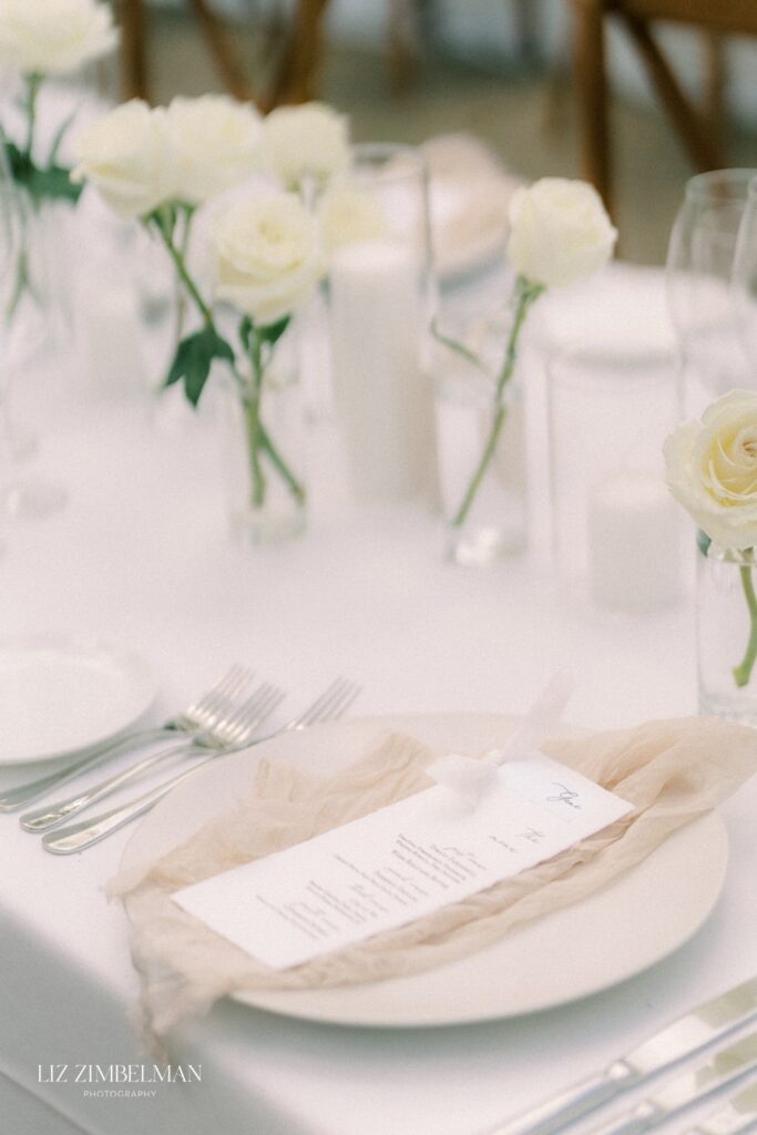 A wedding table setting with white roses, candles, cutlery, and a menu placed on a white plate with a beige napkin.