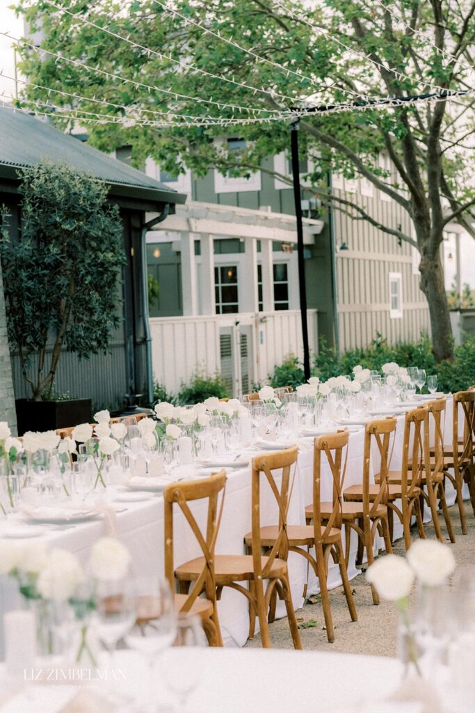 Outdoor dining setup with long tables, white tablecloths, wooden chairs, and floral centerpieces under string lights. Trees and buildings in the background.