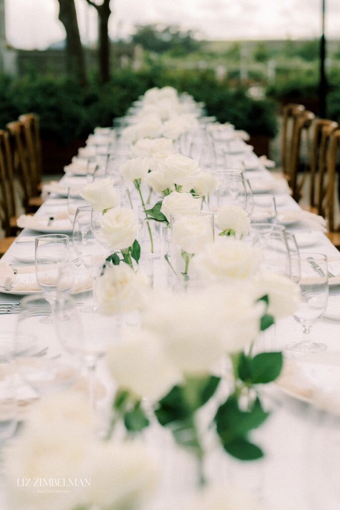 A long dining table is elegantly set with white rose centerpieces, wine glasses, and place settings. The setting is outdoors with blurred greenery in the background.