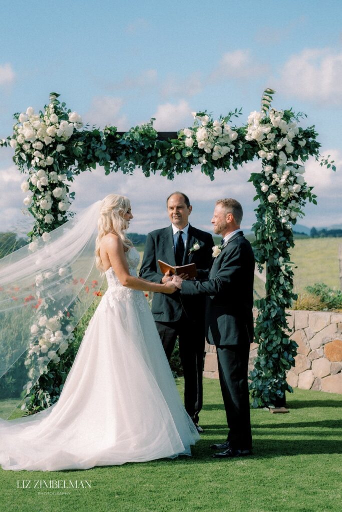 Bride and groom hold hands during an outdoor wedding ceremony under a floral arch, with a person officiating.