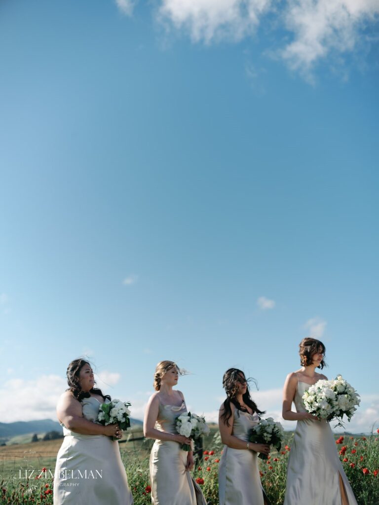 Four bridesmaids in matching light dresses stand outdoors holding white bouquets, with a clear blue sky and fields in the background.