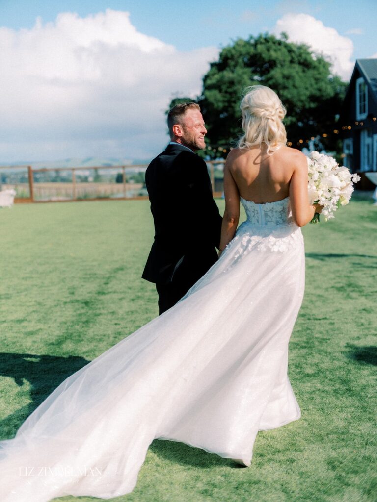 A bride and groom walk across a grassy field. The bride wears a white gown and holds a bouquet. The groom is in a black suit. A building and tree are in the background.