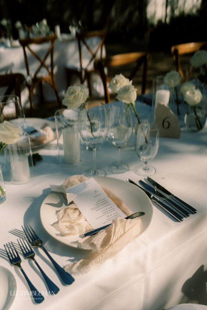 A table setting for a formal event with white plates, silver cutlery, menus, and glassware. White flowers and candles are arranged along the center of the table.