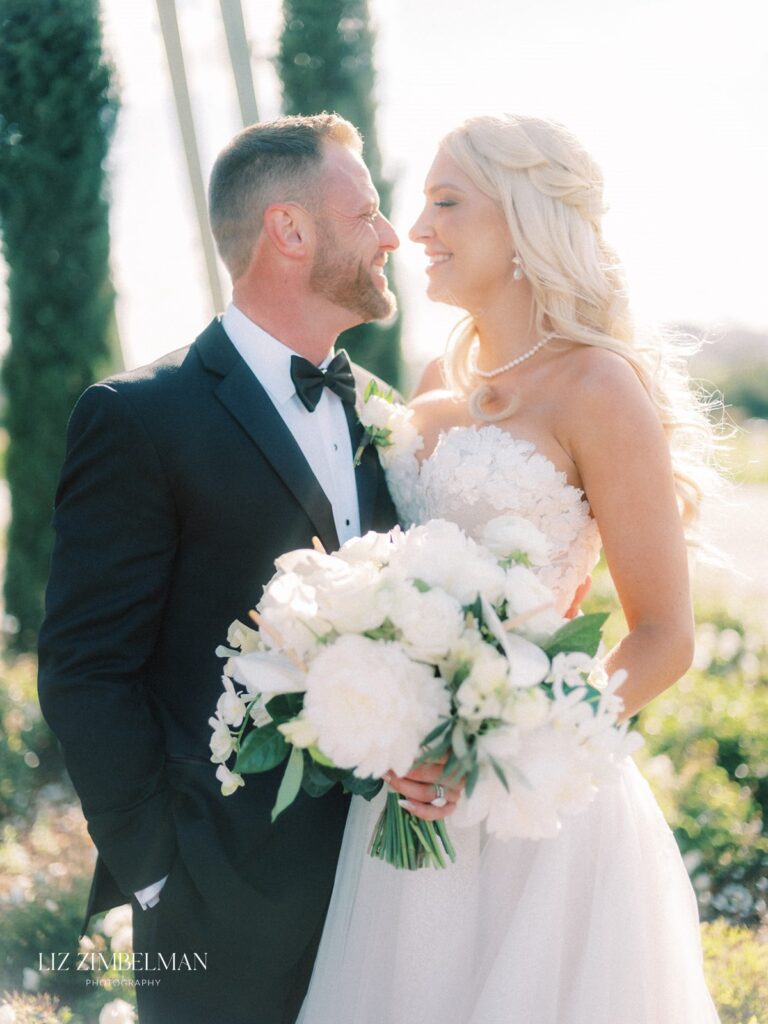 Bride and groom smiling at each other outdoors, bride holding a bouquet of white flowers.