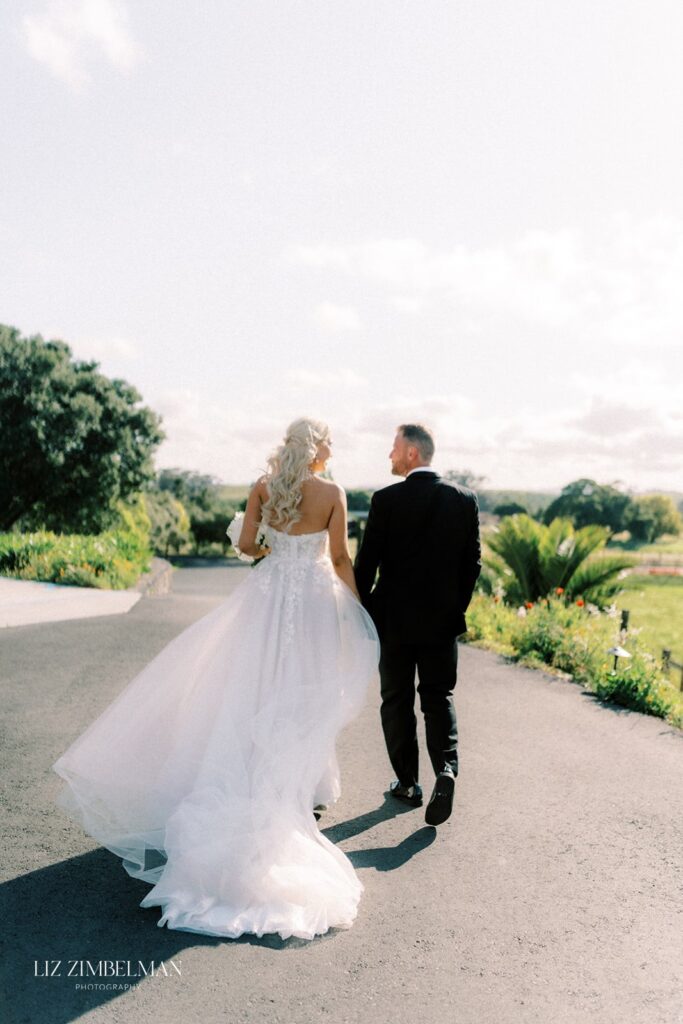 A bride in a white gown and a groom in a black suit walk hand in hand down a sunny outdoor path, surrounded by greenery.