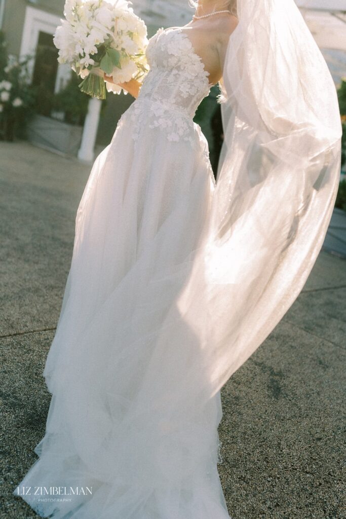 Bride in a white dress holding a bouquet, with a long flowing veil, standing outdoors on a sunny day.