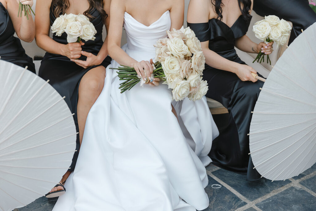 A bride in a white dress sits with bridesmaids in black dresses, holding white roses and parasols.