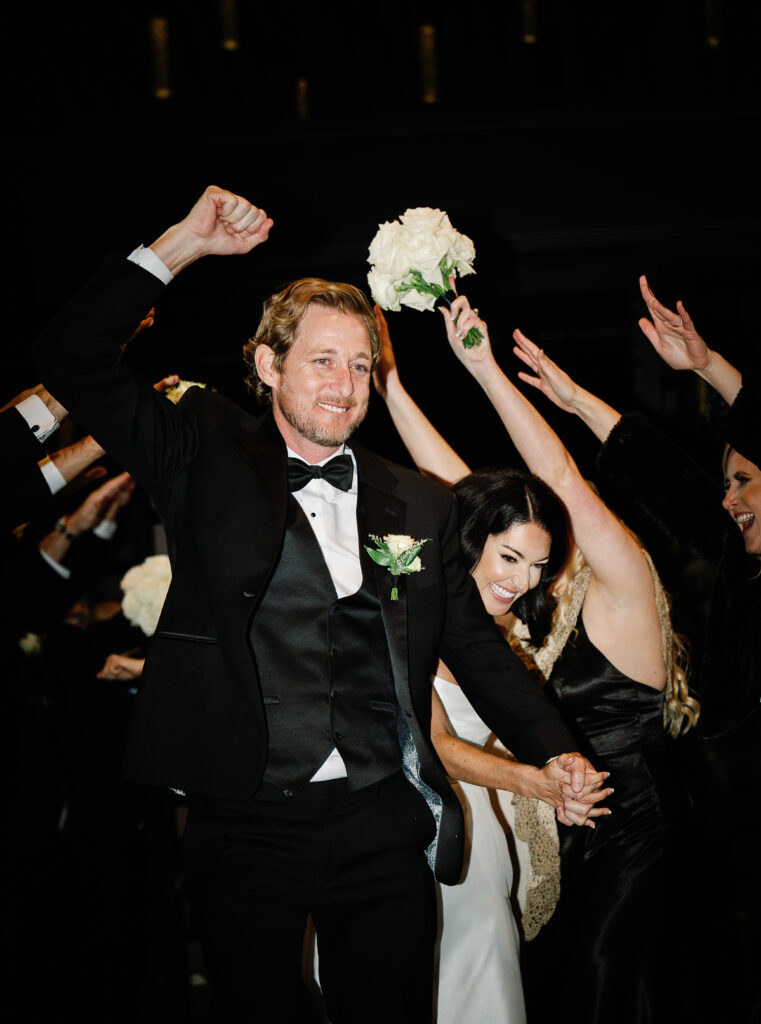 A man in a black tuxedo and a woman in a white dress celebrate with raised arms while walking through a crowd of people dressed formally, some holding white flowers.