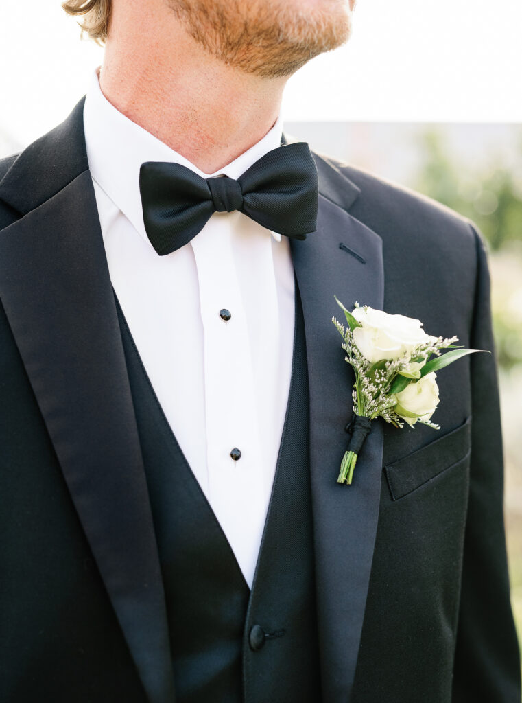 A man in a black tuxedo and bow tie, with a white shirt and black vest, wears a boutonniere of white roses and greenery on his lapel.