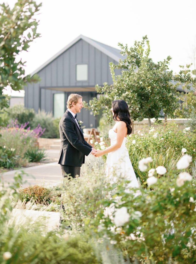A couple stands holding hands in a garden setting, with a modern gray house in the background. The man wears a suit, and the woman is in a white dress.