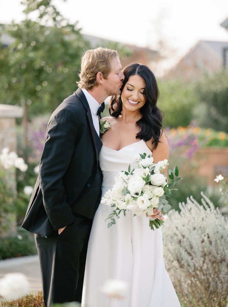 A groom in a tuxedo kisses a bride in a white dress holding a bouquet. They are outdoors with greenery and flowers in the background.