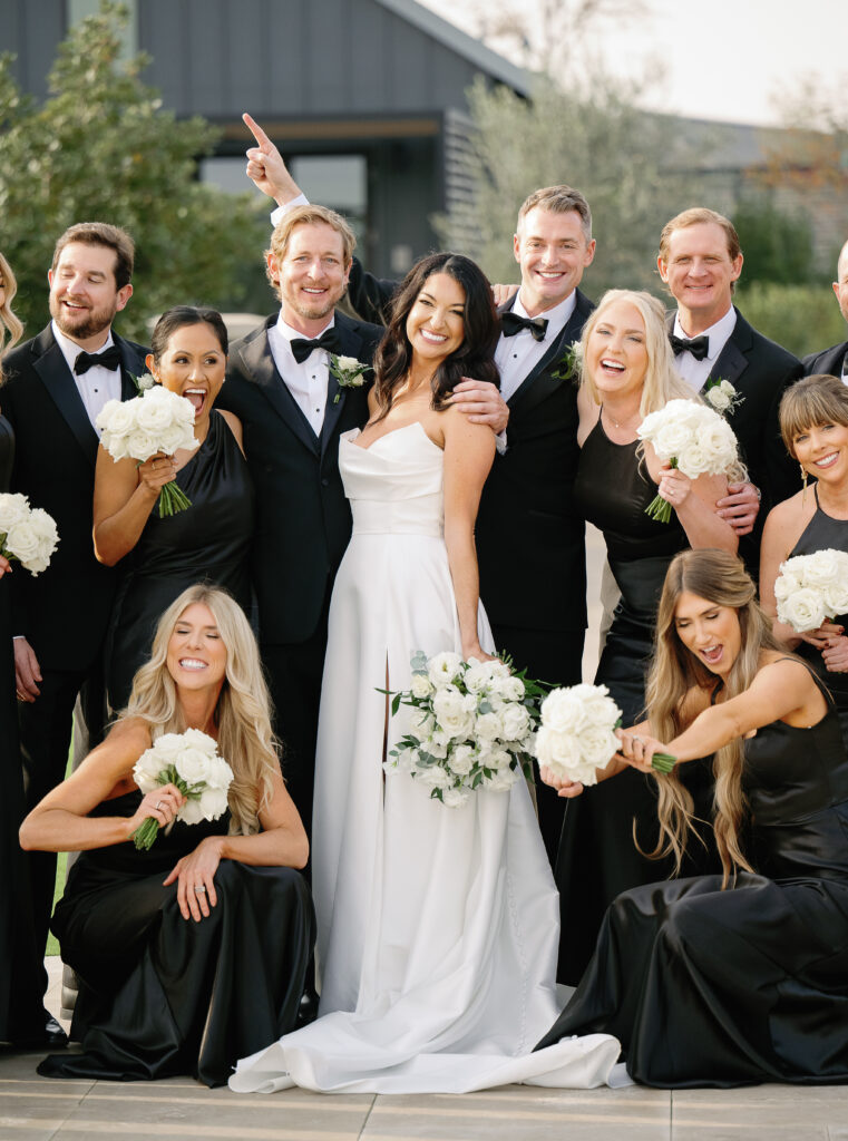 A bride and groom pose with their wedding party. The bride holds a bouquet, and everyone is smiling. The bridesmaids wear black dresses and hold white bouquets.