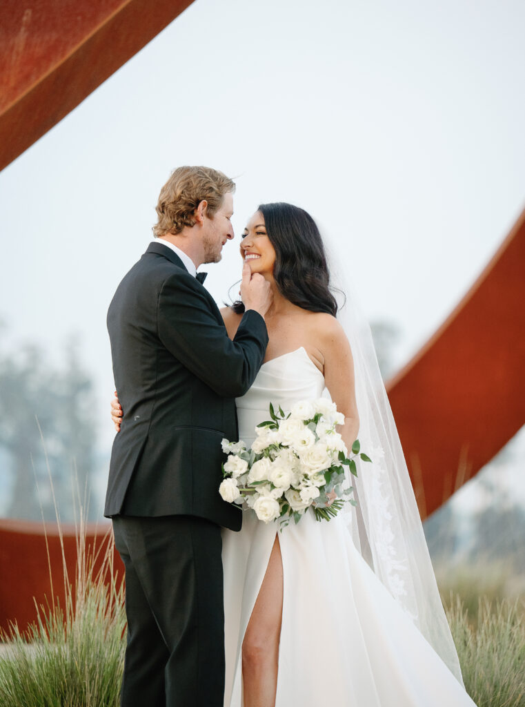 A bride and groom standing close, smiling at each other. The bride holds a bouquet of white flowers. The background shows a large, abstract red sculpture.
