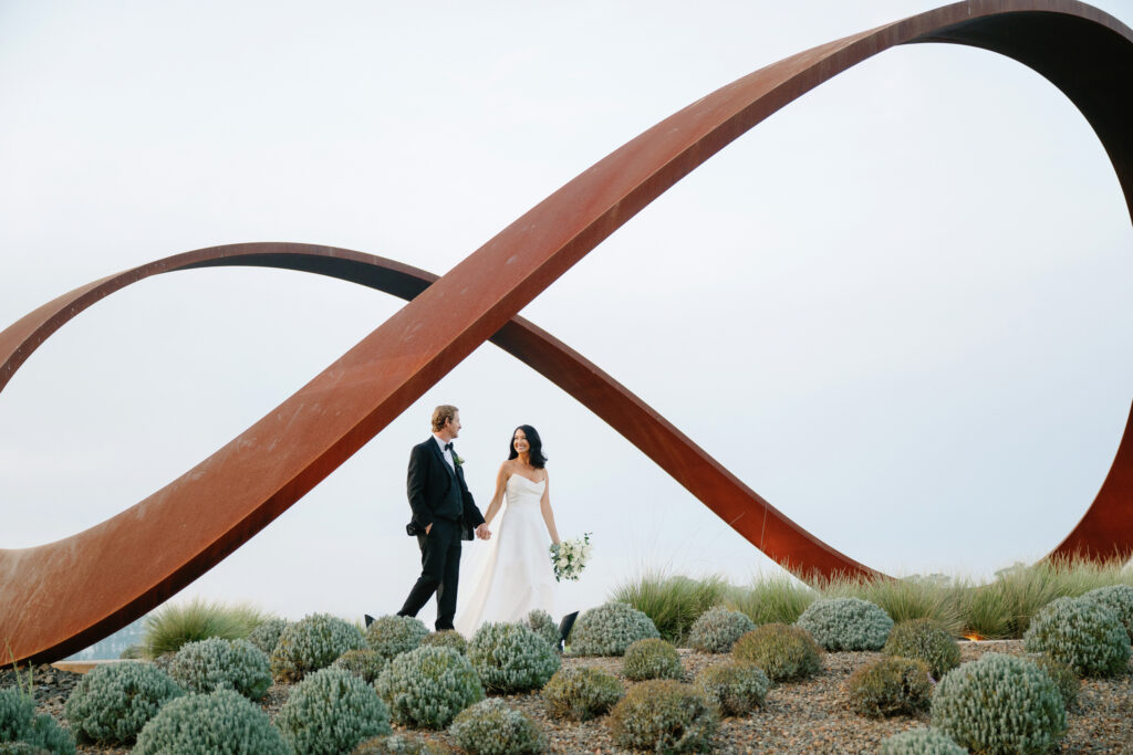 A couple in formal attire holds hands while walking in front of a large, twisting metal sculpture outdoors, embodying the elegance of a Stanly Ranch wedding orchestrated by a renowned Napa wedding planner.