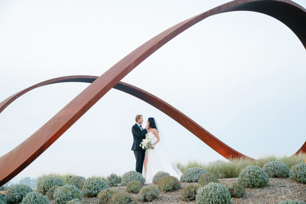 A bride and groom stand facing each other under a large, looping metal sculpture in a landscaped area with low plants at their Stanly Ranch wedding, crafted with elegance by a top Napa wedding planner.