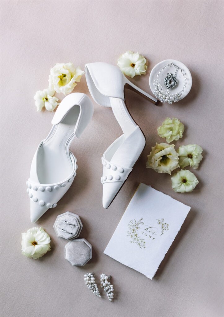 White bridal shoes with pearl details, surrounded by creamy flowers, a vows book, jewelry, and two small boxes on a soft background.