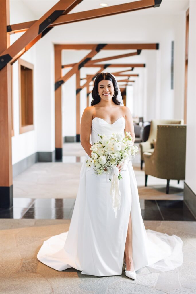 A bride in a white strapless gown holds a bouquet of white flowers while standing in a hallway.