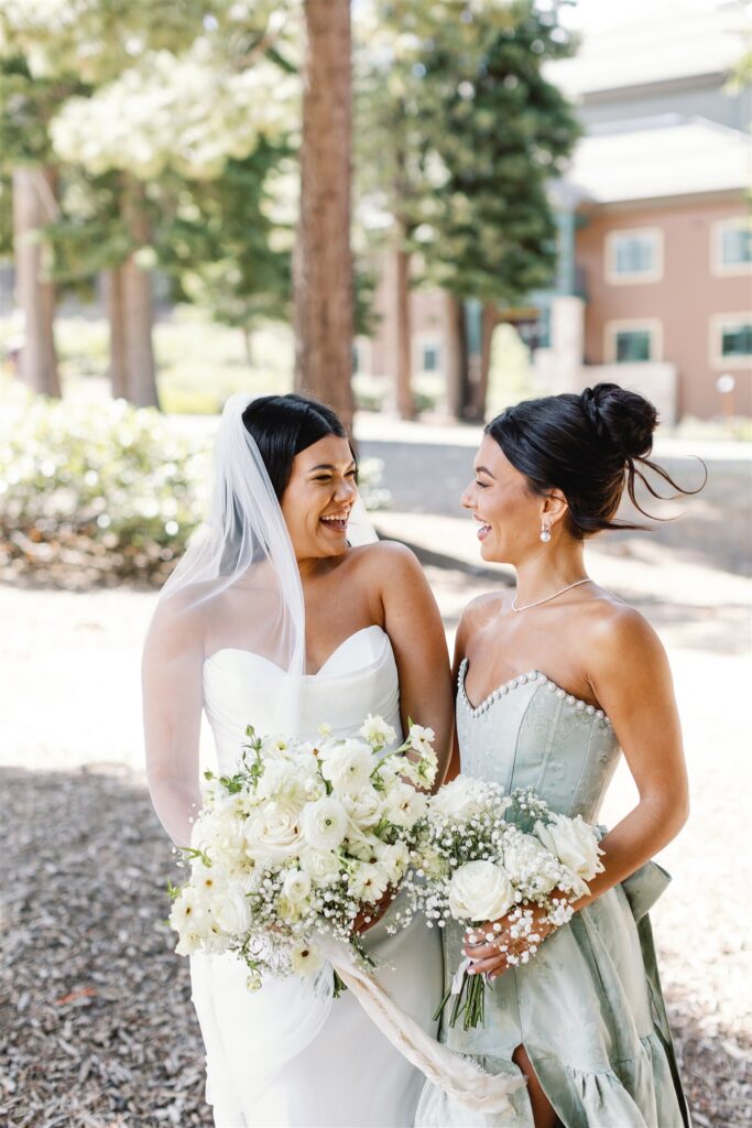 Two women stand outdoors, smiling at each other. One wears a white wedding dress and veil, holding a bouquet of white flowers. The other wears a light green dress, also holding a floral bouquet.
