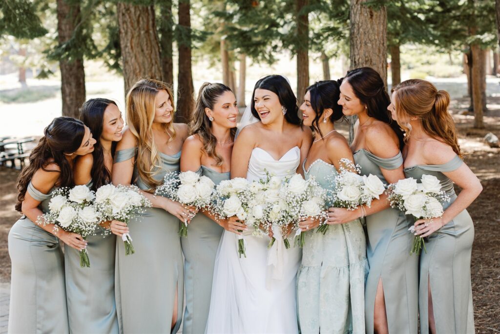 A bride in a white gown stands with six bridesmaids in matching sage dresses, holding bouquets of white flowers, surrounded by trees.