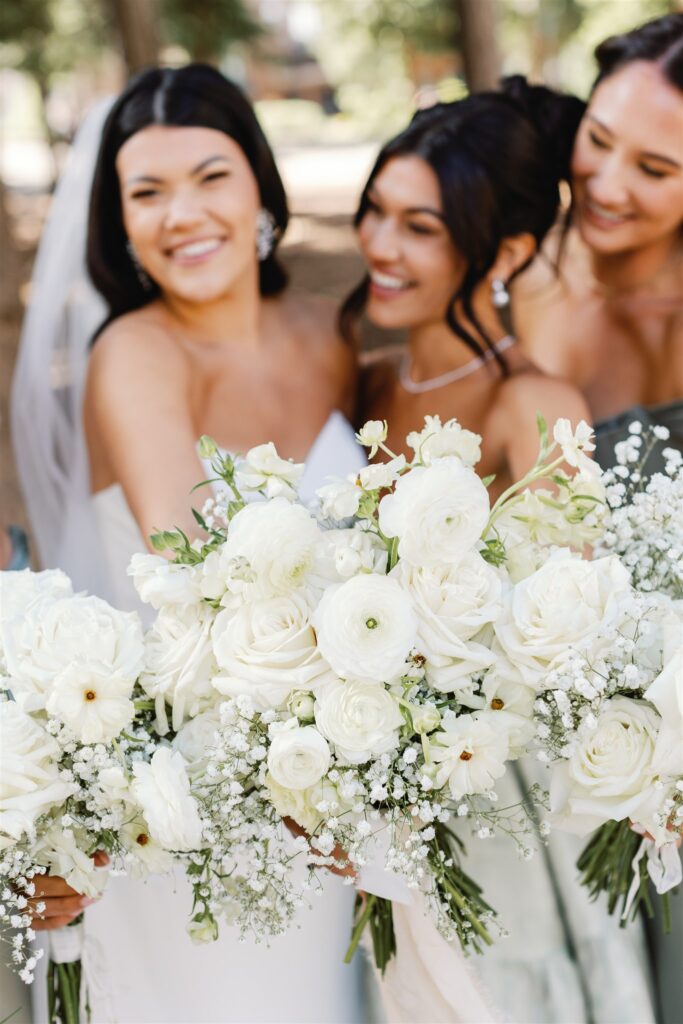 Three women in dresses hold white floral bouquets, smiling in an outdoor setting.