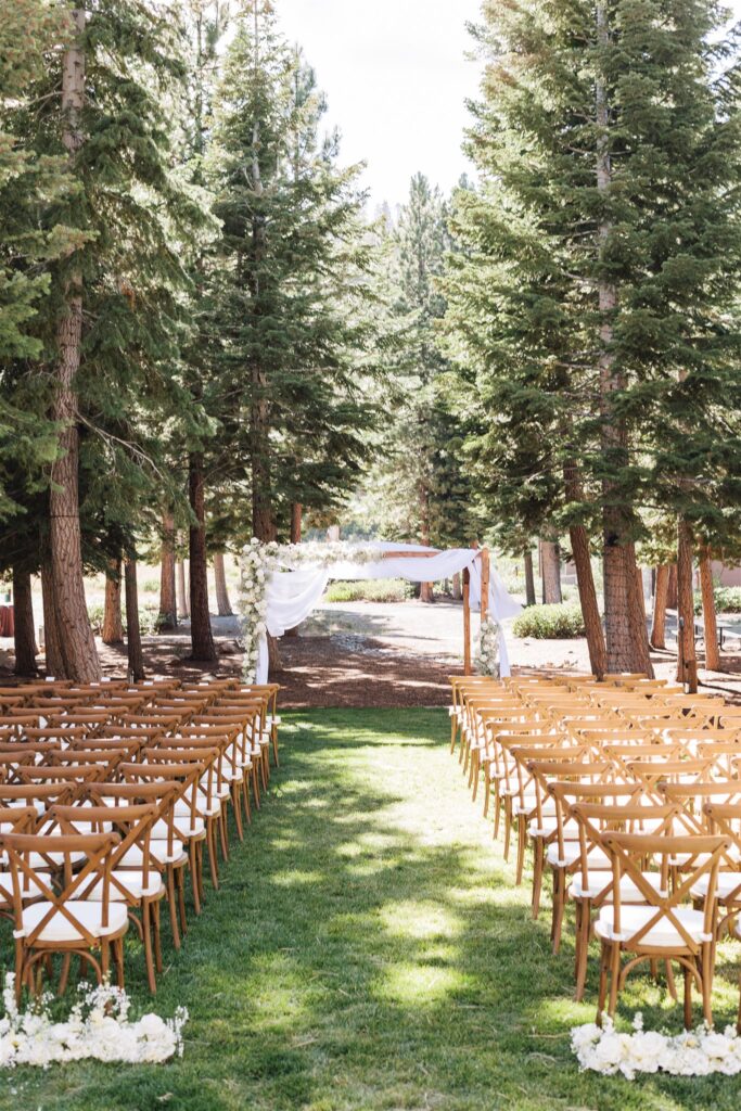 Outdoor wedding setup with wooden chairs lining an aisle leading to a floral arch among tall trees.