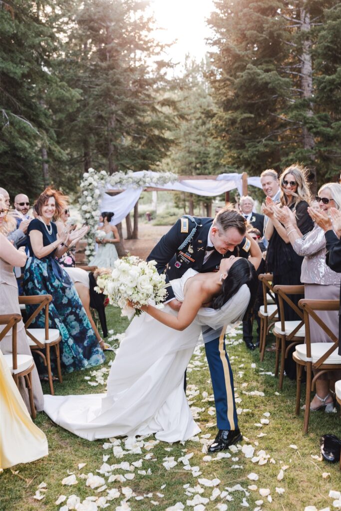 A bride and groom share a kiss at an outdoor wedding ceremony surrounded by guests, with petals scattered on the ground and an archway in the background.