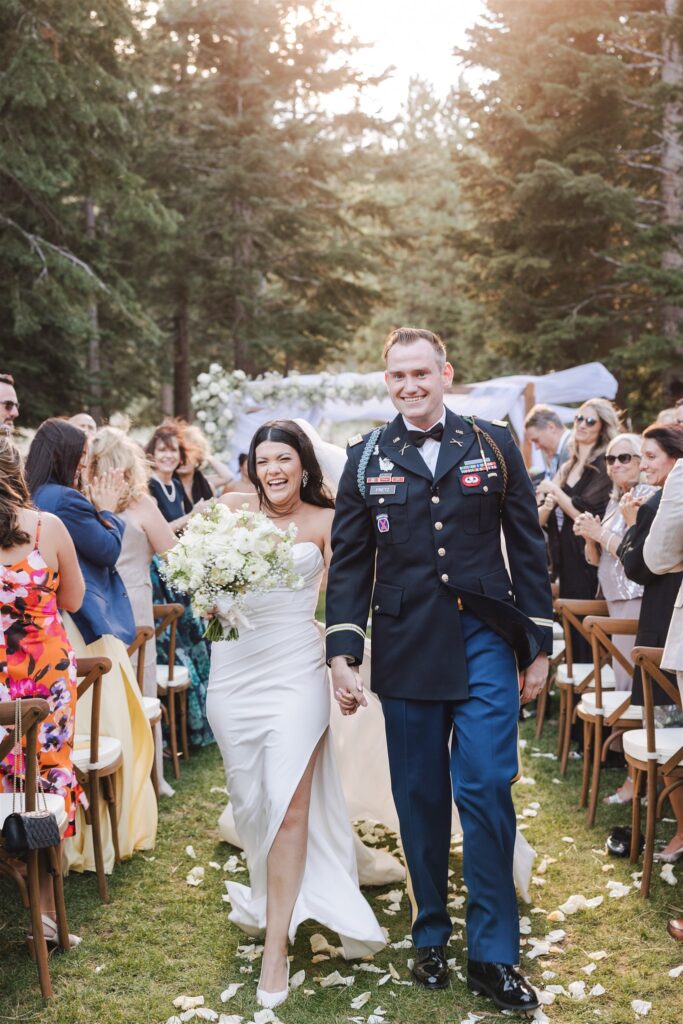 Bride and groom walking down the aisle outdoors, smiling, surrounded by guests and trees.