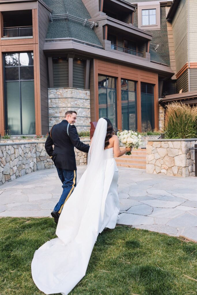 A couple walks hand in hand outside a building; the bride wears a white gown and veil, holding a bouquet, and the groom is in a military uniform.
