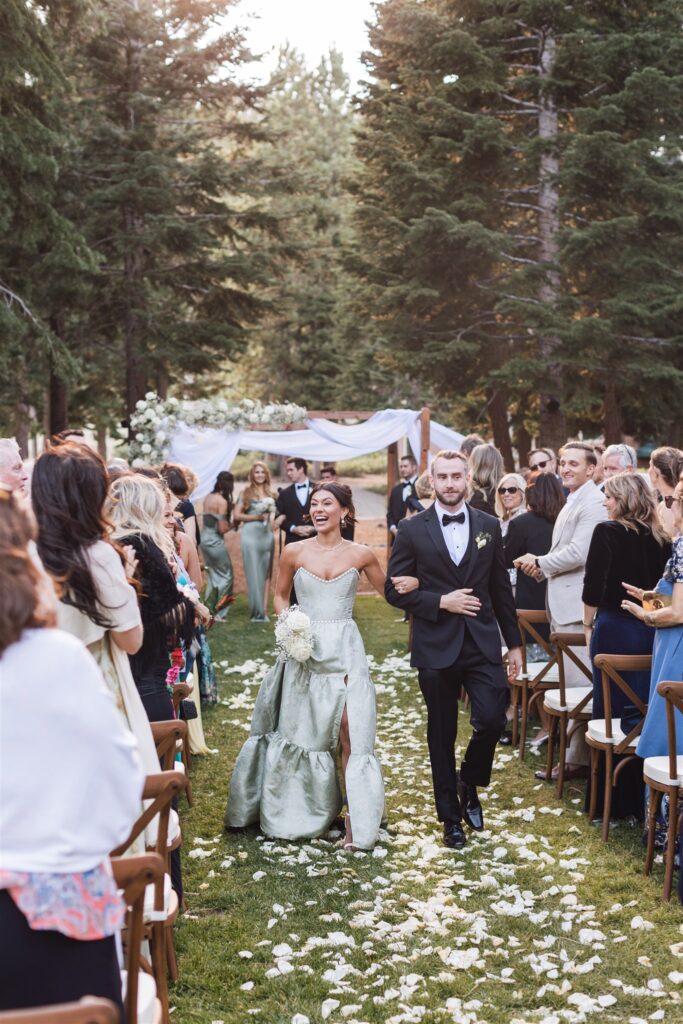 A bride and groom walk down an outdoor aisle lined with guests. The bride wears a green dress, and the groom is in a black tuxedo. They seem joyful. Trees and floral decorations surround the scene.