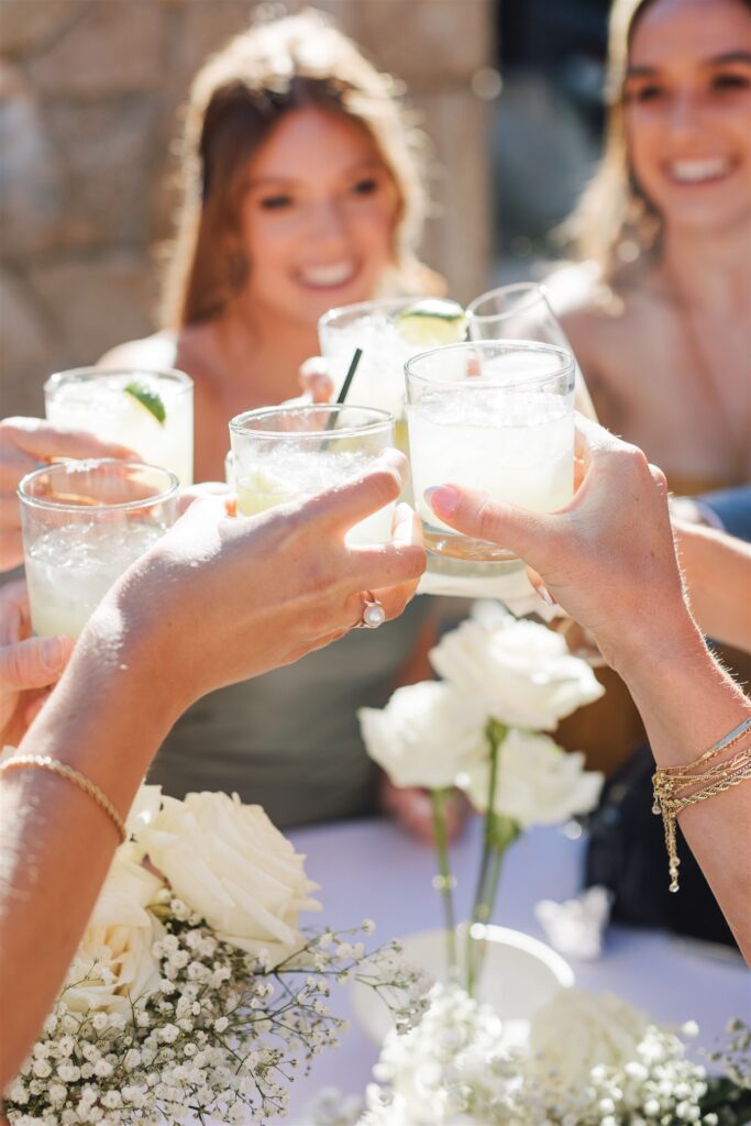 People clinking glasses in a toast, with white flowers on a table and a stone wall in the background.