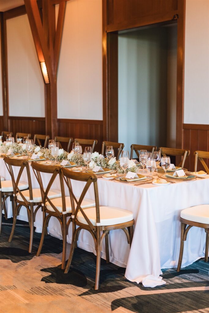 A long dining table is set with white cloth, floral centerpieces, and glassware, surrounded by wooden chairs in a warmly lit room.