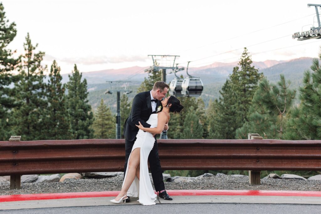 A couple dressed in formal attire shares a dance on a roadside with cable cars and pine trees in the background.