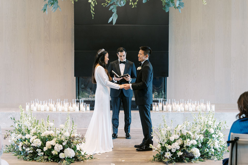 A couple stands holding hands at the altar during their Stanly Ranch wedding ceremony. A person officiates, holding a book. The setting is elegantly adorned with white flowers and numerous candles, creating an atmosphere of timeless romance.