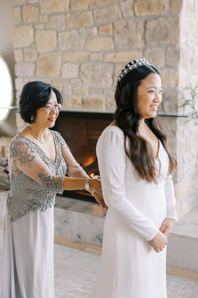 A woman in a white dress is being helped by an older woman in a decorated gown, standing in front of a stone fireplace.