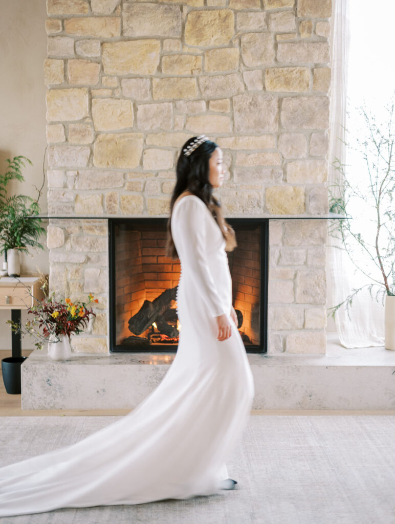 A person in a long white dress walks across a room with a stone fireplace in the background.