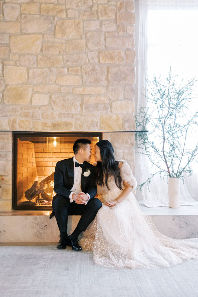 A couple in formal attire sits by a stone fireplace, leaning in close, with a vase of greenery nearby.