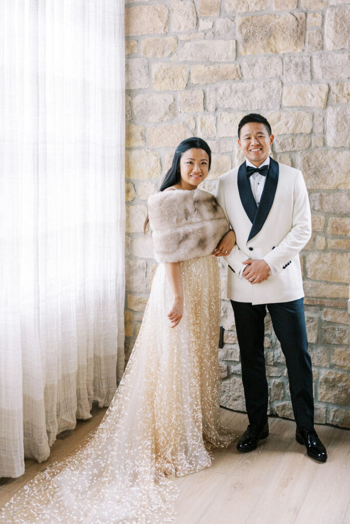 Couple posing indoors; woman in a sparkly gown with a fur stole and man in a white tuxedo jacket and black pants, standing against a stone wall next to a sheer curtained window.