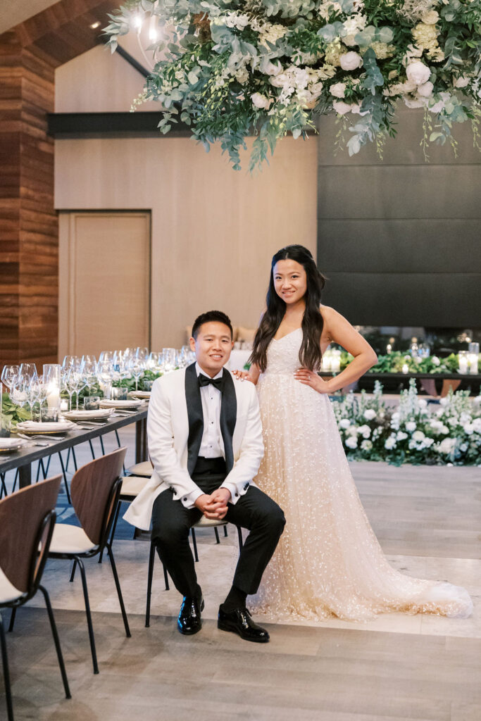 A couple in formal attire poses in an elegantly decorated room with a table set for an event.