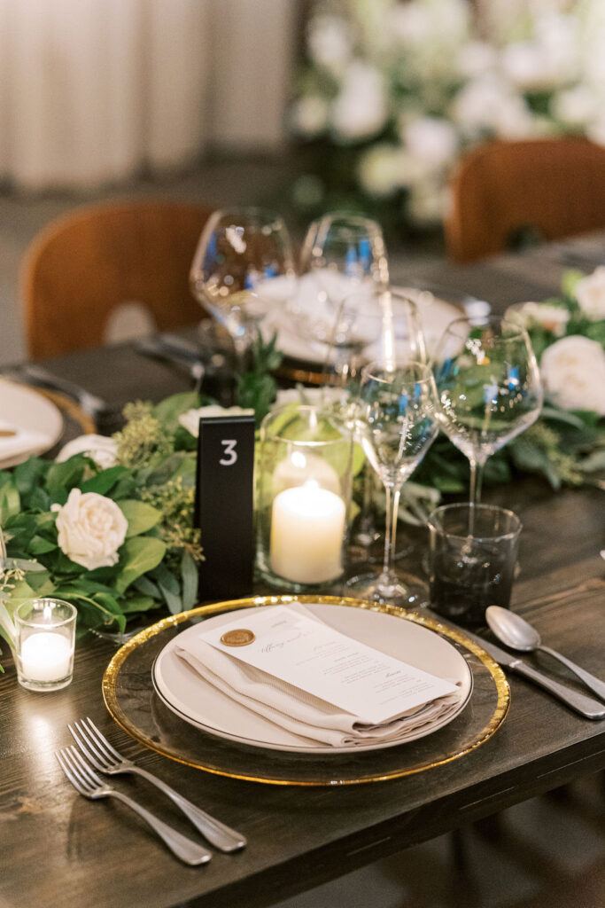 Elegant table setting with a plate, napkin, menu, and cutlery on a dark wooden table. Wine glasses, candles, and greenery adorn the table. A black table number "3" is visible.