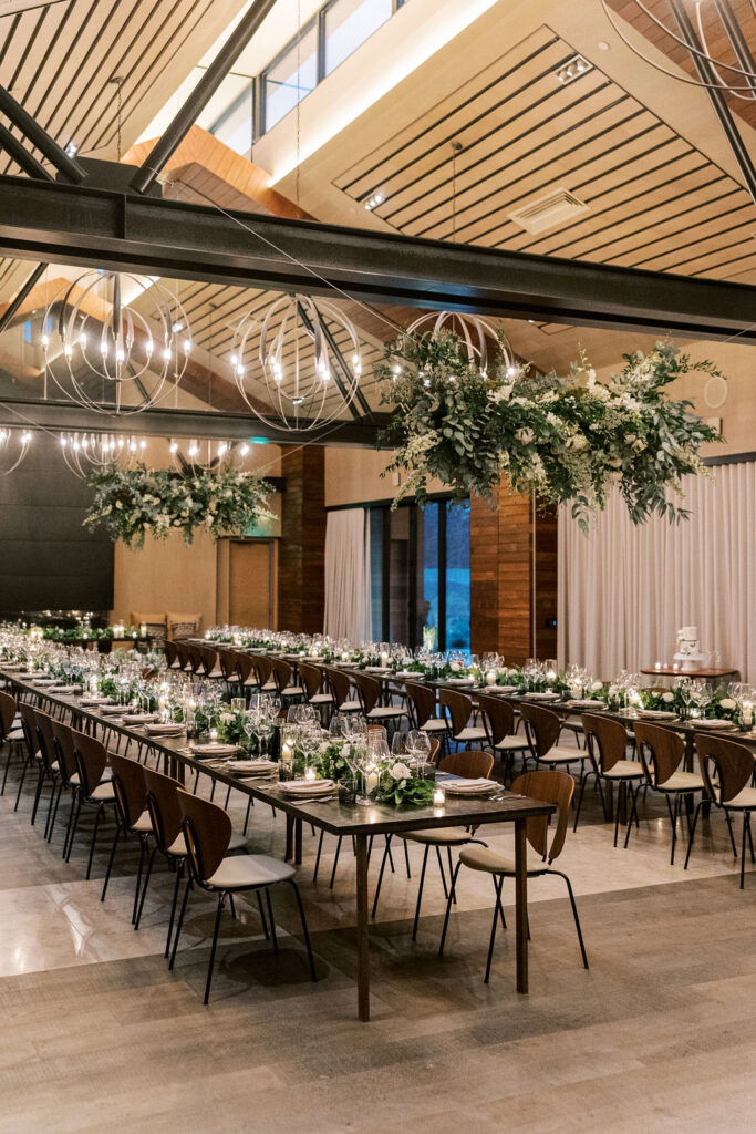 Elegant dining hall with long tables set for an event, featuring hanging greenery and modern spherical chandeliers.