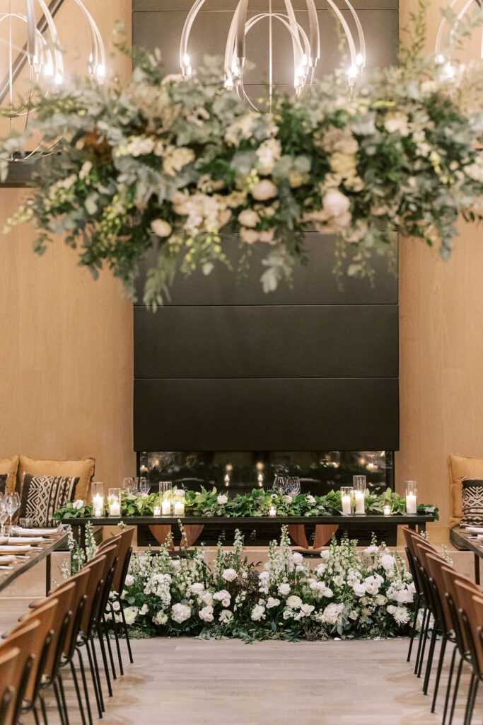 Elegant indoor wedding setting with a floral arch above a table, surrounded by candles and white flowers, against a black wall backdrop. Seats arranged facing the table.
