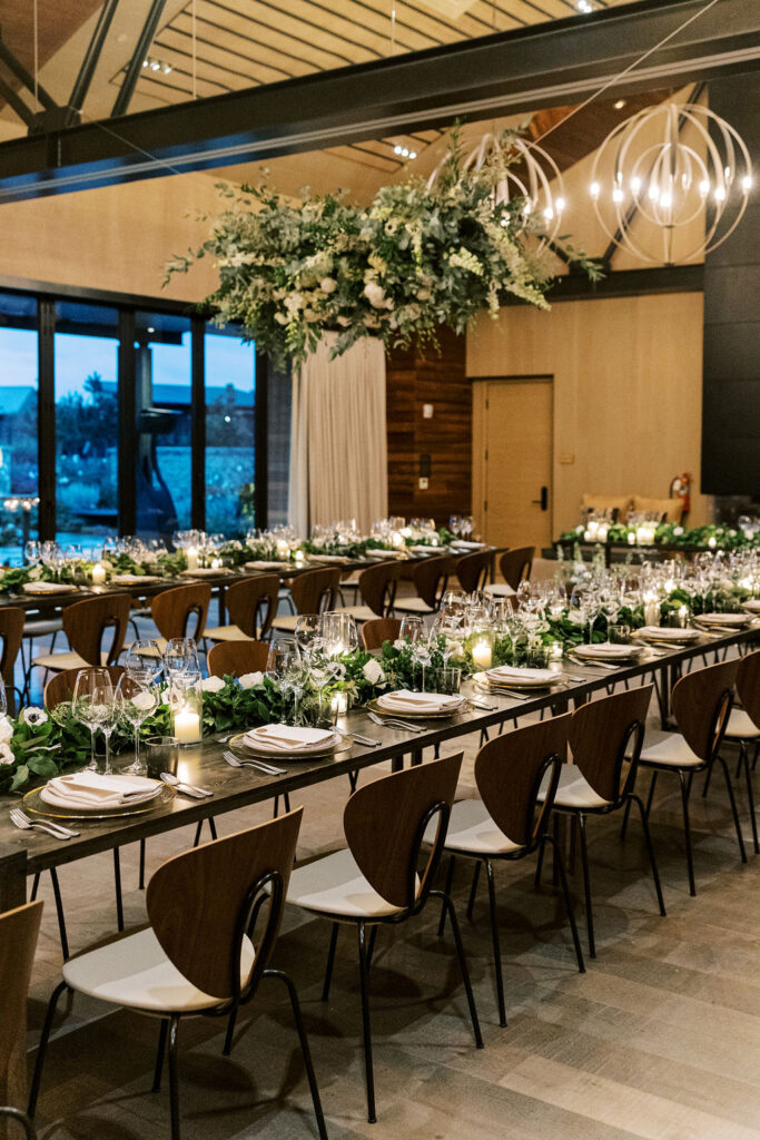 Elegantly set dining tables with glassware, plates, and greenery centerpieces under modern chandeliers in a spacious room.