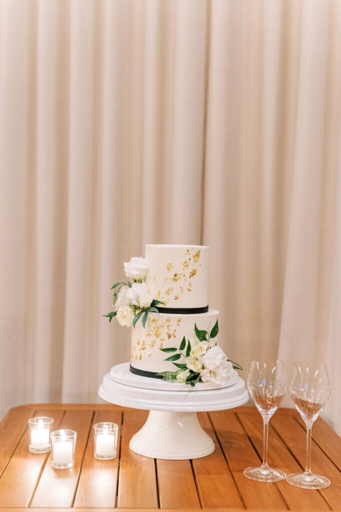 Two-tier white cake with gold accents and white flowers on a stand. Three lit candles and two empty wine glasses are on a wooden table, set against a beige curtain backdrop.