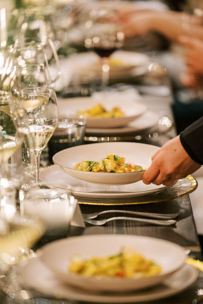 A hand places a dish of pasta on a elegantly set dining table, surrounded by plates and wine glasses.