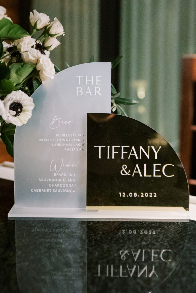 A wedding bar sign with selected beer and wine offerings beside white flowers. The names "Tiffany & Alec" and date "12.08.2022" are displayed.