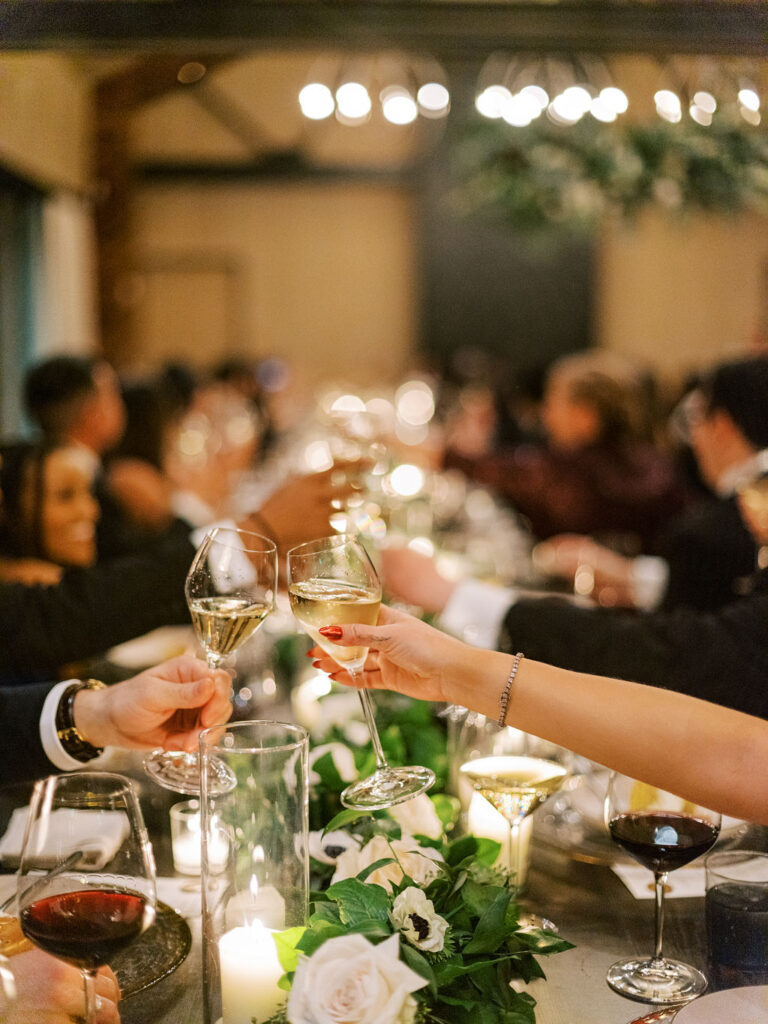 People clinking wine glasses at a long table adorned with flowers and candles in a warmly lit room.